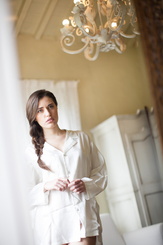 an italian bride looking at herself in the mirror wearing a white silk shirt during her bridal preparation in a rustic hotel room at urbino resort in italy
