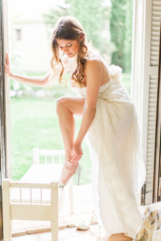 an elegant and fine art wedding portrait of an italian bride getting ready in her rustic bridal suite while she puts on her wedding shoes by the window facing the wedding church in italy