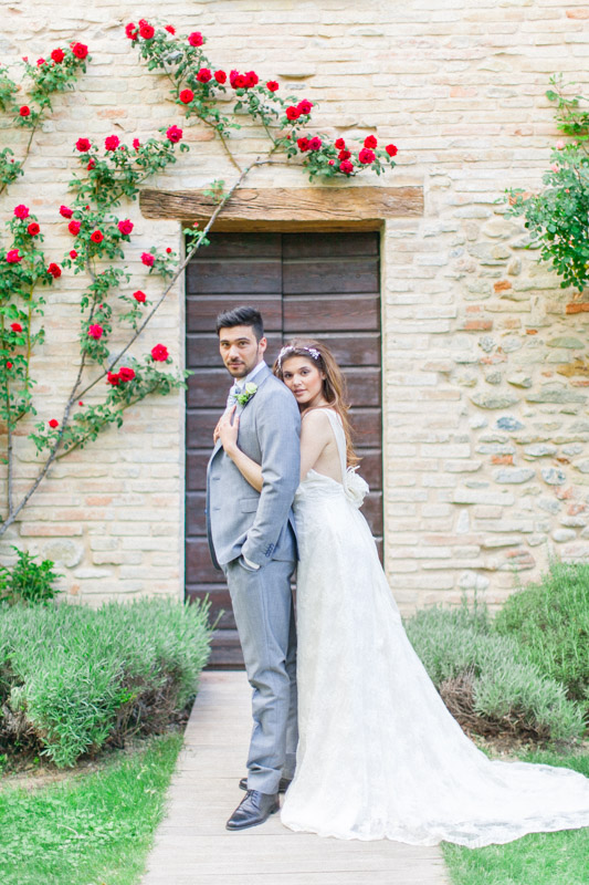 rustic wedding portraits on an italian bride and groom in front of a rustic door with red roses at urbino resort in the marche region in italy