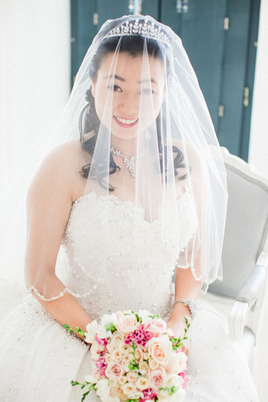 a fine art portrait of a chinese bride sitting on a chair in her bridal suite while she smiles at the camera covered in her bridal veil and wedding crown and holding her wedding bouquet
