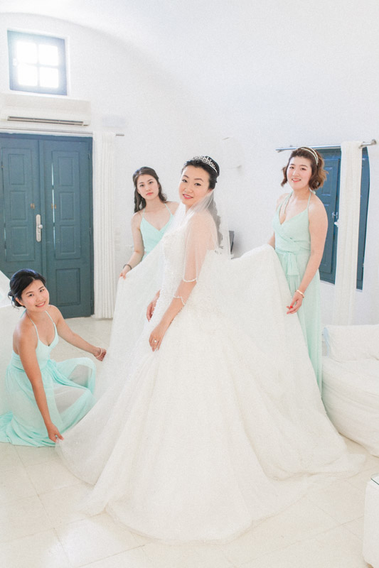 a portrait of a chinese bride and her bridesmaids while they help the bride fixing her wedding dress before the wedding ceremony at sun rocks hotel in santorini