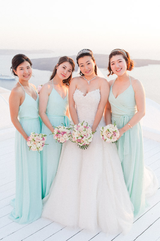 a chinese bride and her chinese bridemaids holding pink and white bouquets after a wedding ceremony at the sun rocks hotel in santorini while wearing light blue bridesmaids dresses