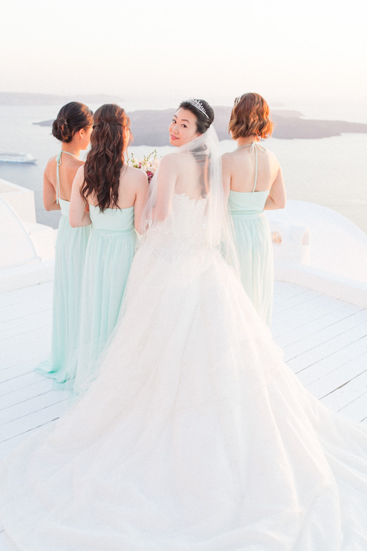 portrait of a bride and her bridesmaids wearing light blue dresses while looking at the breaktaking caldera view in santorini and the bride is turned to look at the photographer