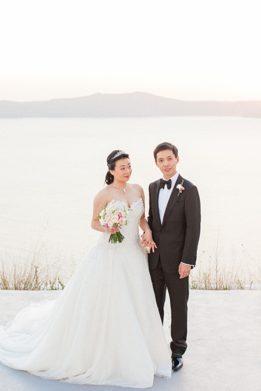 portrait of a chinese bride and groom at sunset time while holding hands after their intimate wedding ceremony at the sun rocks hotel in santorini with a breathtaking caldera view