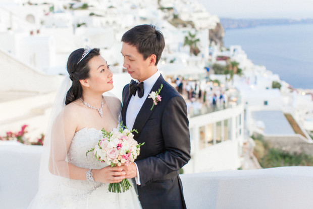 a chinese bride and groom looking at each other at their wedding portraits while the bride holds her wedding bouquet taken in the village of firostefani in santorini